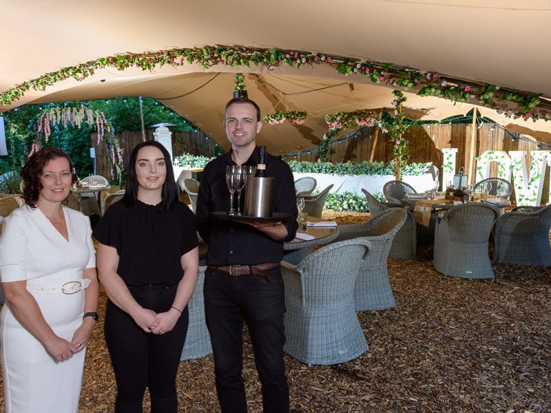 Three people standing under a canopy in front of dinner tables, one of the three is holding a tray with wine bottles