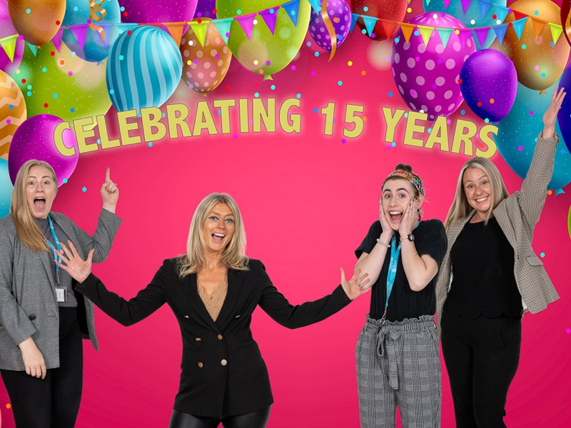 Four people standing under balloons celebrating under the phase "Celebrating 15 years"