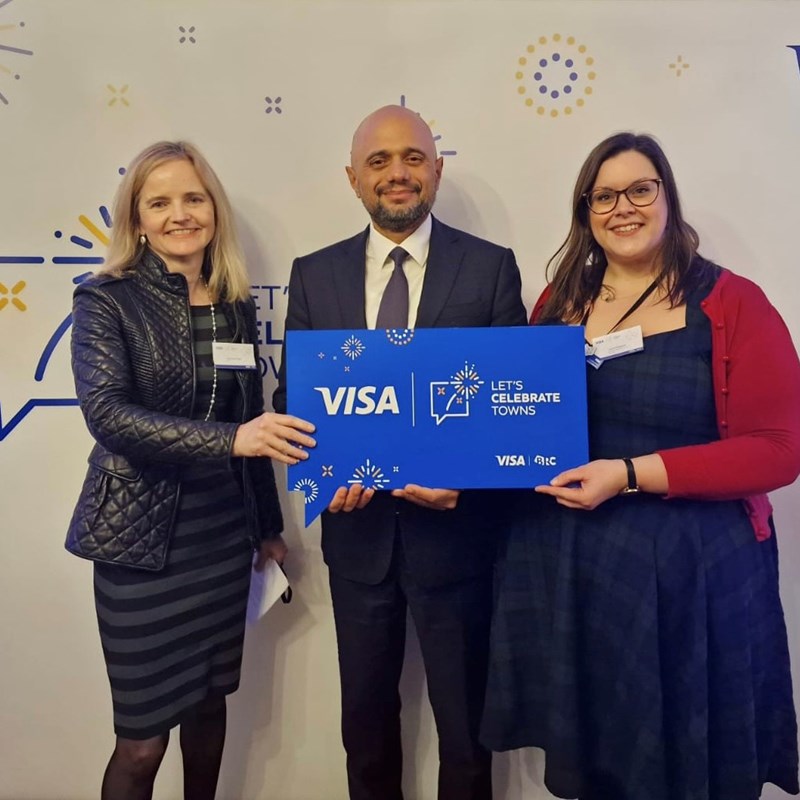 Business women stood either side of a male wearing a smart suit holding a plaque with the text "Let's Celebrate Towns".